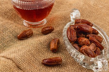 Tarihler, kırsal bir çuval bezi yüzeyinde, koyu bir içecek kadehinin yanına konur ve paylaşmak ve zevk almak için sıcak, davetkar bir ortam yaratır..