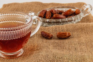 Koyu çay dolu berrak bir bardak, desenli bir çuval bezinin üzerinde, hassas bir hurma tabağının yanında oturur. Ayarlar rahatlamak için ideal bir ortam yaratıyor..