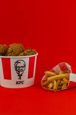 Lviv Ukrayna 05.21.25 Kıtır kıtır kızarmış tavukla dolu bir kova altın kızartma servisinin yanında oturuyor, hepsi canlı kırmızı bir arka plana karşı kurulmuş çarpıcı bir görsel yaratıyor..