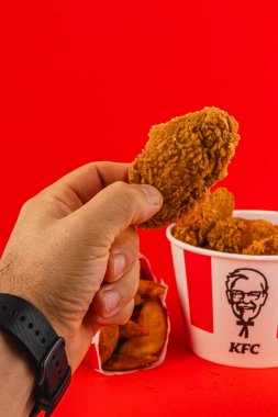 Lviv Ukrayna 05.21.25 Bir el kızarmış tavuk bir parça tutarken arka planda bir kova daha tavuk görünüyor, hepsi parlak kırmızı arka planda..