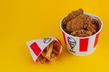 Lviv Ukrayna 05.21.25 Kıtır kıtır kızarmış tavukla dolu bir kap parlak sarı arka planda altın patates kızartmasının yanında oturur. Düzenek, sıradan bir yemek atmosferini vurguluyor..