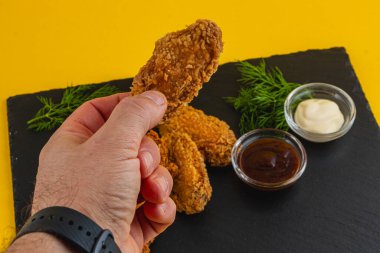 Bir el, kızarmış tavuk kanadını daha fazla kanat ve sos içeren siyah bir tabağın üzerinde tutar. Taze bitkiler sunuma renk katar.