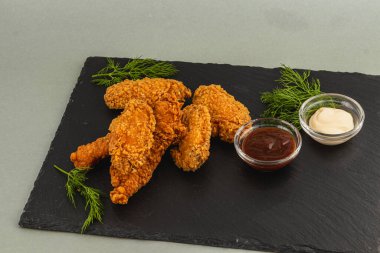 Bir porsiyon kızarmış tavuk, siyah tahta üzerine dizilmiş, yanında iki küçük kase lezzetli soslar ve garnitür için taze otlar var..