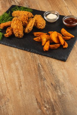 Bir tabakta kızarmış tavuk kanatları ve yanında da sade bir yemek için mükemmel, ahşap bir masada altın tatlı patates dilimleri ve soslar var..