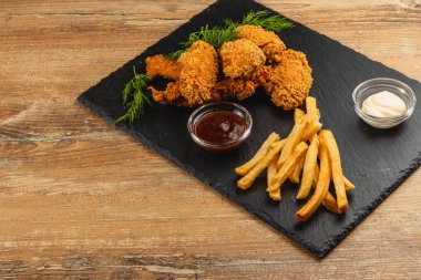 Kızarmış tavuk parçaları siyah bir tabakta, yanında altın kızartmalar ve küçük bir kase sos ve mayonez ile ayarlanarak cezbedici bir yemek oluşturulur..