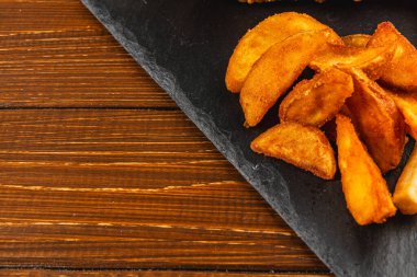 Çıtır patatesler, kalın ahşap bir yüzeye yerleştirilmiş, dokulu siyah bir tabağa yerleştirilir. Lezzetli atıştırmalıklar paylaşmak için davetkar bir ortam yaratır..