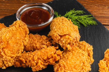 Koyu bir tabakta kızarmış tavuk parçaları, yanında küçük bir kase keskin sos ve garnitür olarak taze dereotu..