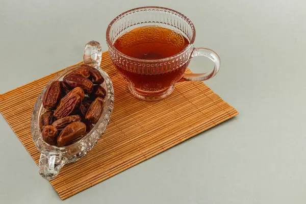 Temiz bir fincan çay bambu bir paspasın üzerinde taze tarihlerle dolu cam bir tabağın yanında oturur. Bu kurulum geleneksel bir atıştırmalık anı sergiliyor, sıcaklığı ve tatlılığı davet ediyor..