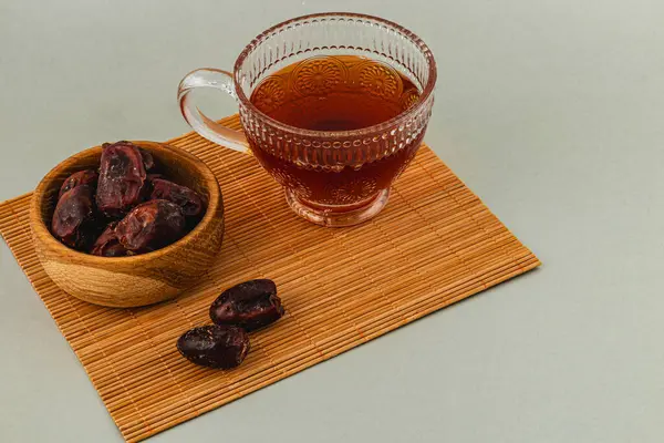 Bir fincan kehribar çayı, zengin ve koyu renkli hurmalarla dolu bir kasenin yanında durur. Kurulum bambu hasırı üzerine kurulmuş sıcak, davetkar bir atmosfer yaratıyor..