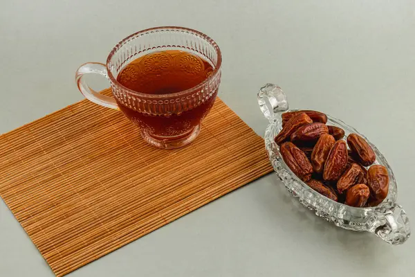 İçi koyu çay dolu hassas bir cam fincana içeride bambu bir minder üzerinde güzelce yerleştirilmiş sulu hurmalardan oluşan kristal bir tabak eşlik eder..