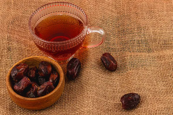 Bir fincan dumanı tüten çay, arka planda hurma dolu bir kasenin yanında oturur. Bu düzenleme samimi ve davetkar bir ortam yaratır..