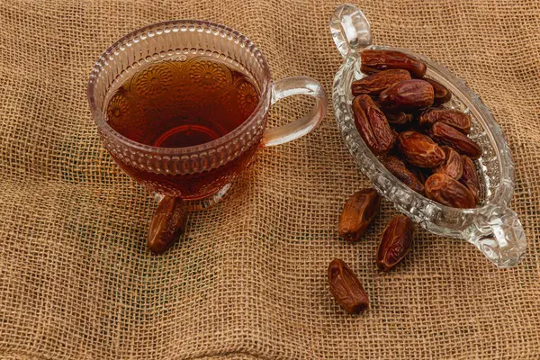 Koyu çayla dolu cam bir bardak çuval bezi yüzeyinde narin bir hurma tabağının yanında oturur. Sahne basit bir hoşgörü anını yakalar..