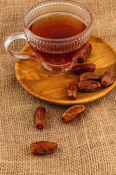 Cam bardakta, lezzetli taze hurma dolu ahşap bir tabağın yanında dumanı tüten kahverengi çay bulunur. Hepsi de kırsal bir arka plana yerleştirilir ve davetkar bir atmosfer yaratır..