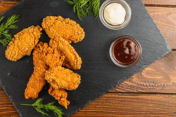 Bir tabakta kremalı ve keskin soslu, sade bir ahşap masada taze otlarla süslenmiş altın kahverengi tavuk kanatları yer alıyor..