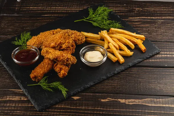 Kızarmış tavuk şeritleri, kızarmış patates kızartması ve iki sosla birlikte, siyah servis tahtasının üzerinde oturur, davetkar ve tatmin edici bir yemek oluşturur..