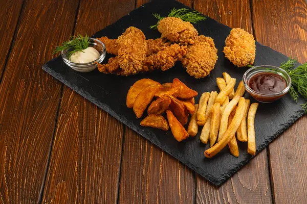 Lezzetli bir tabakta kızartılmış tavuk parçaları ve yanında taze otlarla süslenmiş baharatlı patates parçaları yer alıyor. Baharatlı soslar yemeği tamamlar..