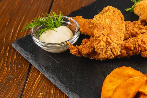 Kızarmış tavuk ihaleleri, yanında küçük bir kase kremalı sos ve taze otlarla süslenmiş siyah bir tabakta servis edilir..