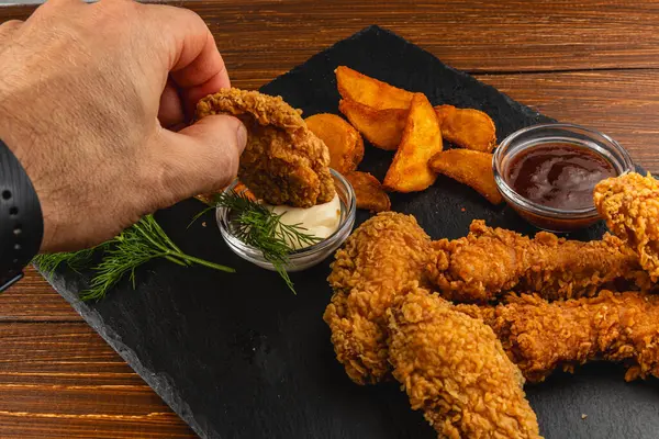 Bir el kızarmış tavuğu kremalı sosa batırıyor. Tabağa altın patates dilimleri ve koyu bir yüzeye batırılmış soslar eşlik ediyor..