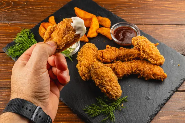 Bir el, yanında patates kızartması ve sade ahşap bir masaya sosla birlikte bir parça kızarmış tavuğa uzanıyor..