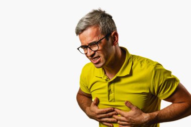 Gözlüklü bir adam, acı belirtileri gösteren karnını tutar. Düz bir arkaplanda duruyor, bir sıkıntı ya da rahatsızlık anının göstergesi..