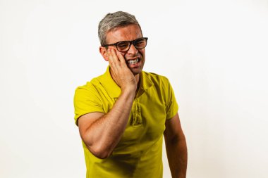 Sarı tişörtlü orta yaşlı bir adam acı dolu bir ifadeyle ayakta duruyor, yüzünü rahatsızlık içinde tutuyor. Geçmişi basit, diş ağrısıyla mücadelesini vurguluyor..