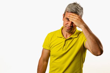 Sarı polo tişörtlü orta yaşlı bir adam, eli alnında hayal kırıklığını ifade eder. Arkaplanda sade bir ışık vardır..