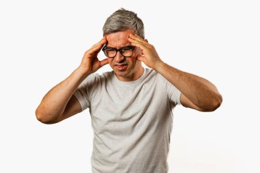 Orta yaşlı, düz gömlekli bir adam iki eliyle şakaklarını tutuyor ve rahatsızlık belirtileri gösteriyor. Tarafsız bir odada, baş ağrısıyla boğuşuyor gibi görünüyor..