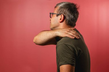 Kısa gri saçlı, gözlüklü bir adam profilde duruyor ve tek eliyle bir stüdyoda canlı kırmızı bir arka planı olan boynuna masaj yapıyor..