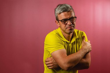 Sarı gömlekli bir adam pembe arka planlı bir stüdyoda duruyor, kaşlarını çatarken kendini kucaklarken rahatsızlık belirtileri gösteriyor..