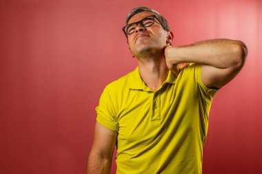 Parlak sarı tişörtlü bir adam kırmızı arka planda duruyor, boynunu acılı bir ifadeyle nazikçe ovuyor, rahatsızlık ya da gerilimi gösteriyor..
