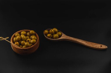 Yeşil zeytinlerle dolu ahşap bir kase daha fazla zeytin içeren bir tahta kaşığın yanında oturur. Koyu arka plan, kâse ve zeytinin doğal tonlarını vurgular..