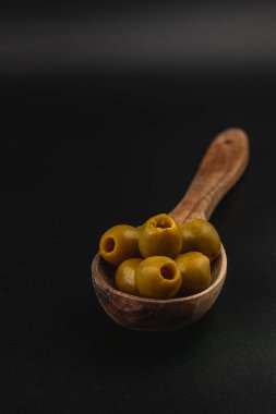 Ahşap bir kaşık birkaç yeşil zeytin içerir, pürüzsüz dokularını ve zıt bir koyu arka plana karşı zengin renklerini sergiler. Sunumun basitliği onların doğal güzelliklerini vurguluyor..