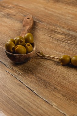 Yeşil zeytinler ahşap bir kaşıkta ve bir şişin üzerinde kırsal bir ahşap masada güzelce sergilenir. Bu ayar zeytinlerin doğal dokularını ve toprak tonlarını vurgular..