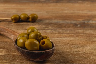 Yeşil zeytinler ahşap bir kaşıkta dizilir, pürüzsüz dokuları ve doğal parıltıları sergilenir. Kırsal ahşap arka plan sahnenin organik hissini tamamlıyor..