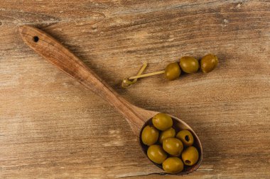 Ahşap bir kaşık birkaç yeşil zeytin tutarken zeytinli bir şiş ahşap masada duruyor. Odunun doğal dokusu ortamı ısıtır, aperatifler için mükemmeldir..