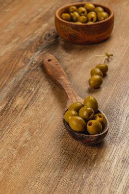 Yeşil zeytinler ahşap bir kaşığa ve kırsal bir ahşap masada bir kaseye konur. Doğal ışık sıcak bir atmosfer yaratıyor, odunların ve zeytinlerin dokusunu vurguluyor..