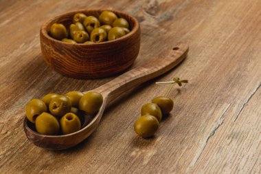 Taze yeşil zeytinler ahşap bir kasenin içinde zeytin dolu bir tahta kaşığın yanında sergilenir. Kırsal masa yüzeyi davetkar düzenine sıcaklık katar. Atıştırmalık ya da aperatif için mükemmeldir..