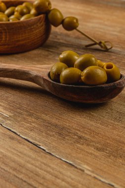 Taze yeşil zeytinler ahşap bir yüzey üzerinde duran ahşap bir kaşığa konur. Arka planda bir kase zeytin görünür, samimi ve davetkar bir atmosfer yaratır..