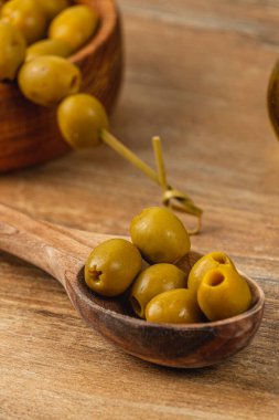 Çeşit çeşit yeşil zeytinler ahşap bir kaşıkta, ahşap bir masanın üzerindeki dekoratif kaselerin yanında, bu lezzetli atıştırmalıkların basit ama davetkar bir sunumunu sergiliyorlar..