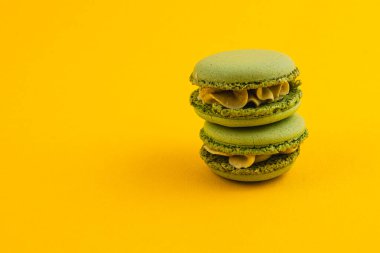 Kremayla dolu iki yeşil badem kurabiyesi canlı sarı bir yüzeye istiflenir, pürüzsüz dokularını ve davetkar renklerini sergiler, onları görsel olarak çekici kılar..