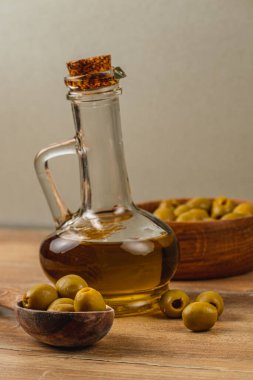 Altın zeytinyağıyla dolu cam bir şişe yeşil zeytinlerle dolu ahşap bir kasenin yanında duruyor. Sahne, sade ahşap bir masa, doğal bir mutfak sahnesi sergiliyor..