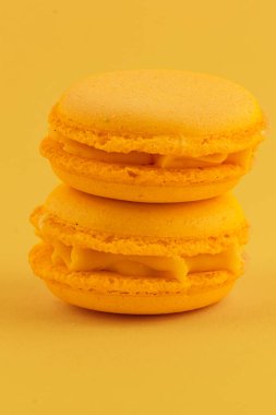 Kremalı iki sarı badem kurabiyesi, parlak sarı bir zemine karşı istiflenmiş. Tatlılar mükemmel pişmiş ve davetkar..