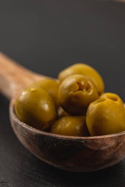 Tahta bir kaşık taze toplanmış ve parlayan canlı yeşil zeytinler taşır. Koyu arkaplan, zengin renklerini ve dokularını ön plana çıkararak, davetkar bir mutfak görüntüsü yaratıyor..