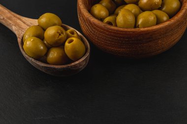 Taze yeşil zeytinler, koyu bir yüzeyde, yuvarlak ahşap bir kasede, ahşap bir kaşıkta dinleniyor, pürüzsüz dokularını ve canlı renklerini vurguluyor..
