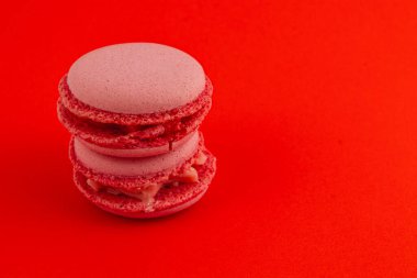 Kremayla dolu iki pembe badem kurabiyesi koyu kırmızı bir zemine karşı yan yana oturur narin dokularını ve çekici renklerini minimalist bir ortamda sergilerler..