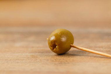 Yeşil bir zeytin bir kürdanla delinir ve kırsal bir ahşap masaya dik konur. Yumuşak doğal ışık, zeytinin pürüzsüz dokusunu ve ayarların basitliğini vurgular..