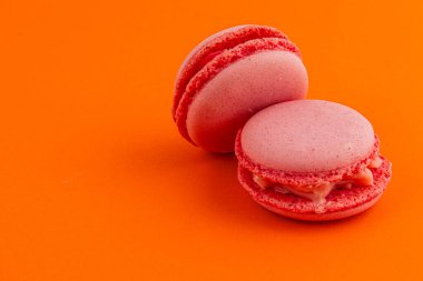 Kremalı iki pembe badem kurabiyesi parlak turuncu bir yüzeye yan yana yerleştirilir ve renkli ve cezbedici bir tatlı görüntüsü yaratır..