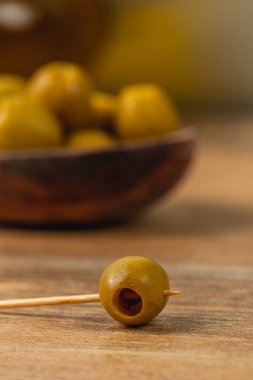 Tek bir yeşil zeytin, tahta bir yüzey üzerinde duran bir kürdanla delinir. Bulanık yeşil zeytinler bir kâseyi dolduruyor, sade ve sade bir atıştırmalık sergiliyor..