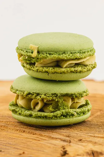 İki canlı yeşil makaron üst üste istiflenmiş, kremalı bir dolgu içeriyor. Hafif ahşap arka plan ikramın cazibesini ve renklerini arttırır.