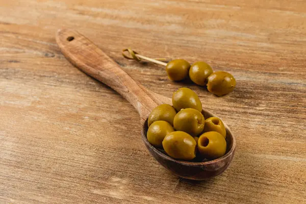 Ahşap bir kaşık yeşil zeytin tutar, parlak yüzeyi ve eşsiz şekilleri sergilenir. Köy tabakası sahneye sıcaklık katarak zeytinlerin doğal cazibesini arttırıyor..